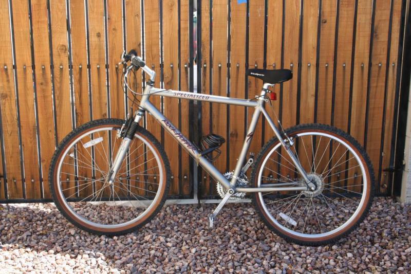 Specialized Rockhopper: A silver mountain bike with thick tires, featuring a black seat and handlebar grips, is leaning against a wooden fence. The bike has distinctive orange accents on the wheels and is positioned on a rocky surface.