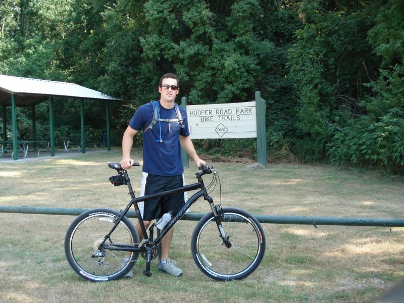 Specialized Rockhopper: A person standing next to a black mountain bike in front of a sign that reads "Hooper Road Park Bike Trails." The individual is wearing a blue t-shirt and black shorts, with a backpack and headphones. There is a picnic shelter in the background surrounded by trees.