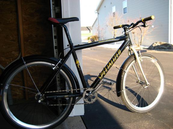 Specialized Rockhopper: A black Specialized bicycle with yellow lettering, featuring a single gear system and wide tires, parked in a garage setting. The background includes a glimpse of a house and a clear driveway.