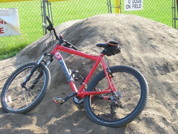 Specialized Rockhopper: Red mountain bike leaning against a sand mound, with a chain link fence and a sign stating "No Dogs on Field" in the background. The scene is outdoors in a grassy area.