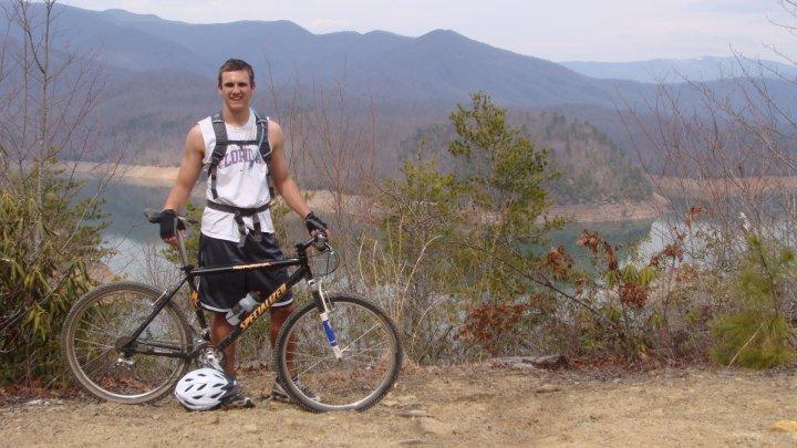 Specialized Rockhopper: A young man stands next to his mountain bike, smiling and posing for the camera. He is wearing a tank top and shorts, along with a helmet resting on the ground. Behind him is a scenic view of a lake surrounded by mountains, with trees and sparse vegetation in the foreground. The sky is partly cloudy, indicating a pleasant day for outdoor activities.
