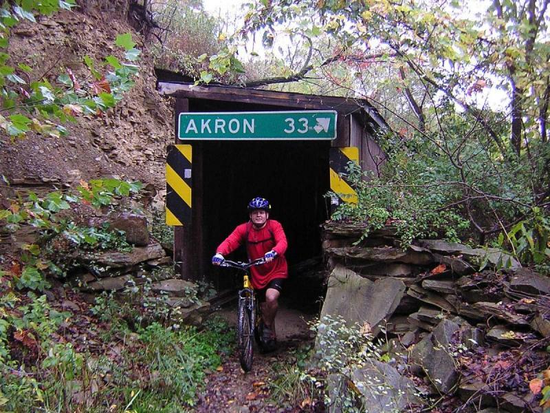Specialized Rockhopper: A mountain biker in a red shirt pushes his bike out of a dark tunnel, with a green sign above indicating "AKRON 33" to the right. The tunnel is surrounded by rocky terrain and overgrown foliage, suggesting a natural setting.