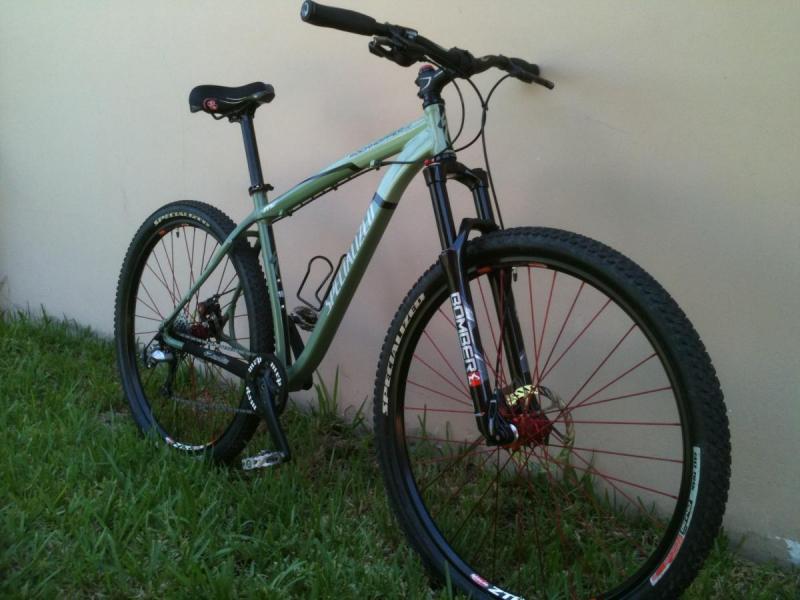 Specialized Rockhopper: A green mountain bike with black accents, featuring a sturdy frame, oversized tires with red spokes, and a front suspension fork, leaning against a light-colored wall with grass in the foreground.