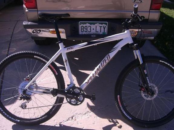 Specialized Rockhopper: A white Specialized mountain bike is parked on a concrete surface, showcasing its sleek design and features. The bike has black components, including the handlebars and wheels, and is fitted with disc brakes and knobby tires. In the background, a gray vehicle and a glimpse of green grass are visible.