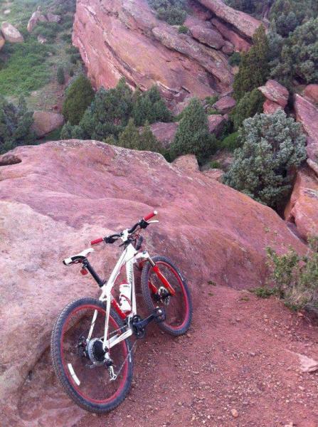 Specialized Rockhopper: A white mountain bike with red accents rests on a rocky outcrop overlooking a lush green valley, surrounded by rugged red rock formations and sparse trees.