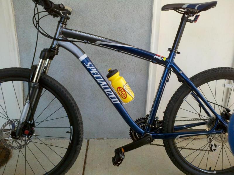 Specialized Rockhopper: A blue mountain bike with a white "Specialized" logo, featuring rugged tires and a front suspension fork. A yellow water bottle is mounted on the frame. The bike is positioned against a light-colored wall with visible flooring beneath it.