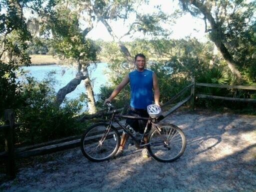 Specialized Rockhopper: A person stands next to a mountain bike on a sandy path by a river, surrounded by greenery and trees. The individual is wearing a blue sleeveless shirt and holding a bike helmet.