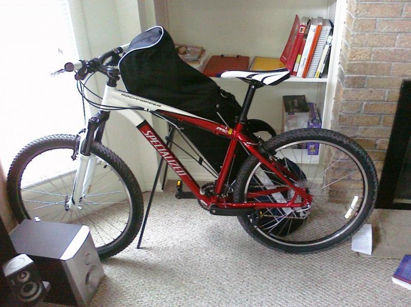 Specialized Rockhopper: A red and white mountain bike leaning against a wall inside a living space, with a black seat cover attached. Nearby, there's a bookshelf filled with colorful books and a speaker, against a backdrop of carpet flooring.