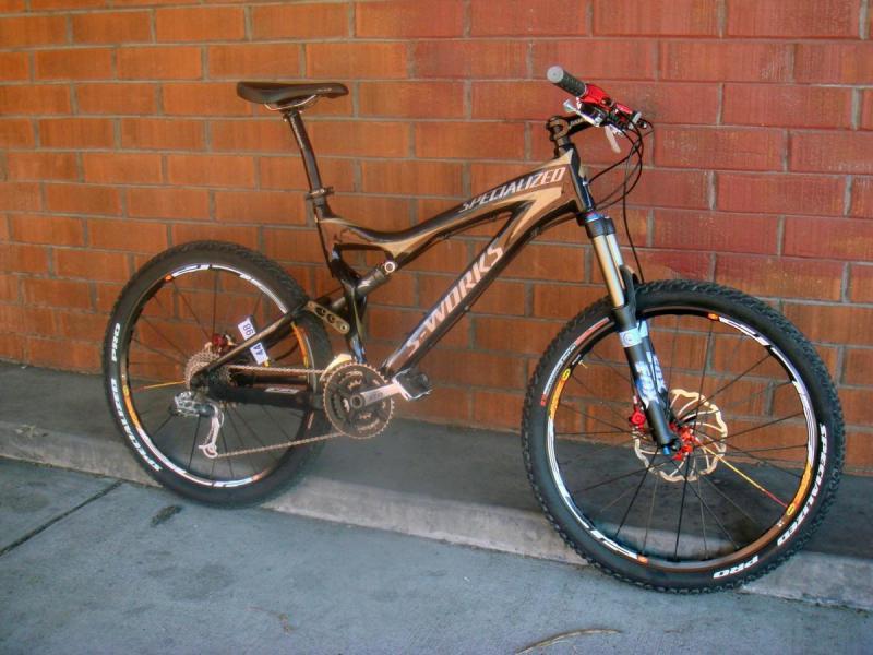 Specialized S-Works Stumpjumper FSR Carbon: A black Specialized S-Works mountain bike leaning against a brick wall. The bike features prominent suspension forks, thick tires, and a sleek frame design. The background consists of a textured red brick wall, and the bike is positioned on a concrete surface.