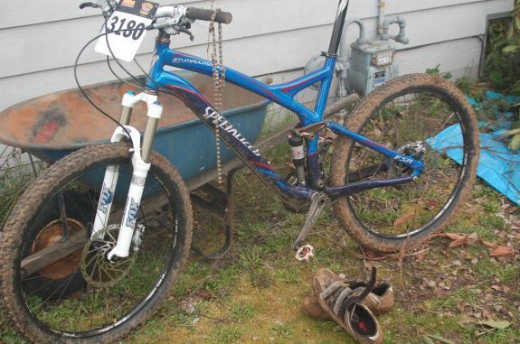 Specialized Stumpjumper Comp: A blue and black mountain bike with muddy tires is leaning against a wooden wall. It features a white front fork and has a race number tag attached to the handlebars. Nearby, a wheelbarrow and a pair of brown shoes are visible. The ground is covered in grass and leaves, suggesting an outdoor setting.