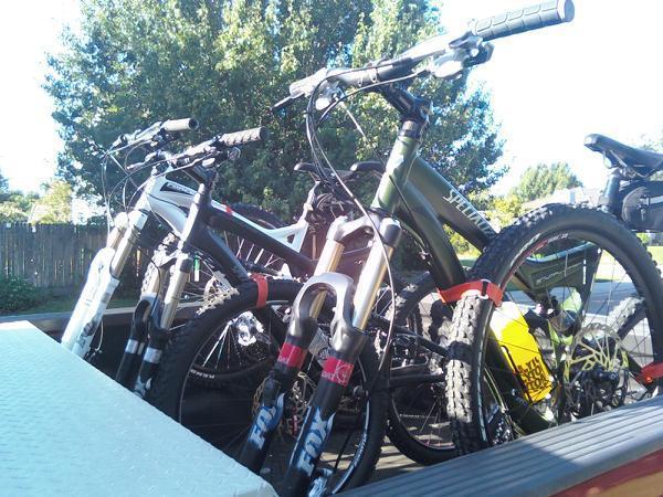 Specialized Stumpjumper Comp: A close-up view of several mountain bikes securely loaded in the back of a truck. The bikes have various colors and designs, showcasing their front suspension systems. In the background, trees and a wooden fence are visible, suggesting an outdoor setting.