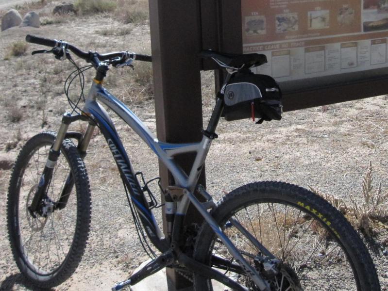 Specialized Stumpjumper Comp: Mountain bike parked next to an informational sign in a desert landscape, with dry vegetation and gravel in the background. The bike features a silver frame with blue accents and thick tires. The sign includes various printed materials, but specific content is not visible.