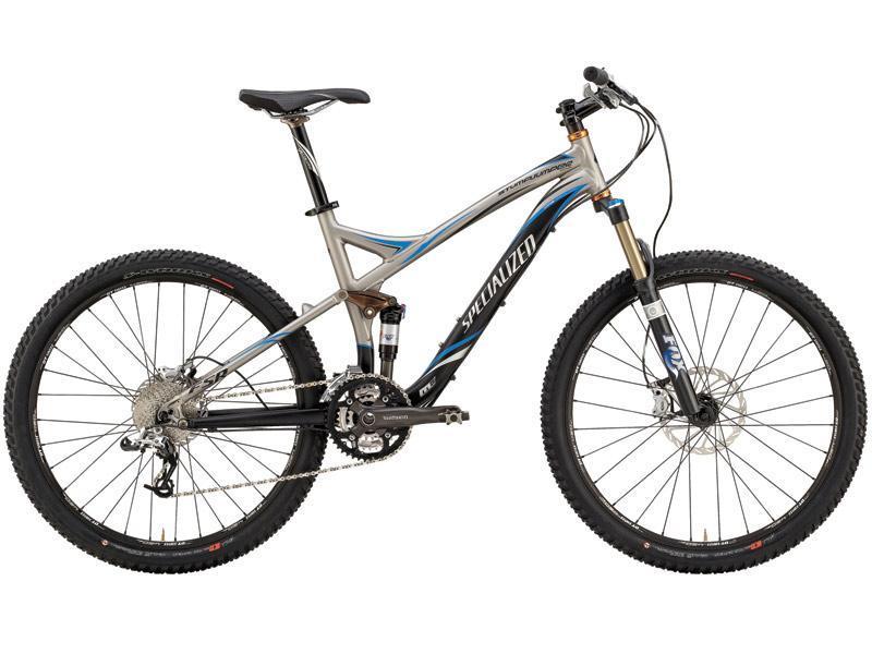 Specialized Stumpjumper Comp: Alt text: A silver and black mountain bike with blue accents, featuring thick tires, a front suspension fork, and a gear drivetrain, positioned on a plain background.