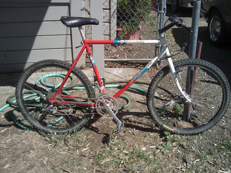 Specialized Stumpjumper Comp: Alt text: A red and white mountain bike is parked against a wall, partially obscured by green garden hoses on the ground. The bike has thick tires and a simple frame design. In the background, a chain-link fence and part of a car are visible.