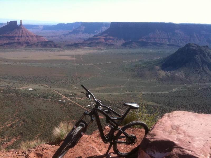 Specialized Stumpjumper FSR: A mountain bike rests on a rocky outcrop with a panoramic view of a vast desert landscape, featuring distant mesas and rugged terrain under a clear blue sky.