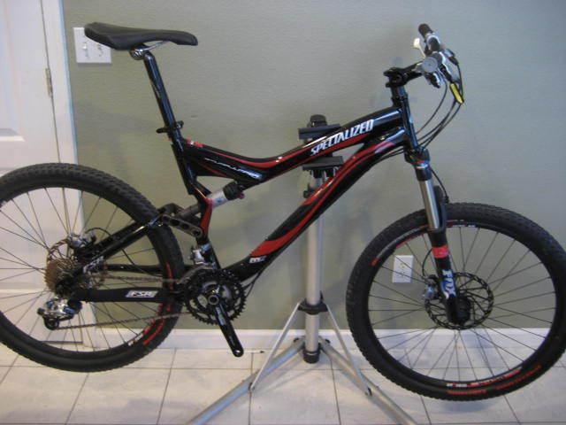 Specialized Stumpjumper FSR: Mountain bike displayed on a repair stand, featuring a black and red frame with the brand name "Specialized" visible. The bike is equipped with knobby tires, a disc brake system, and multiple gears, set against a plain green wall.