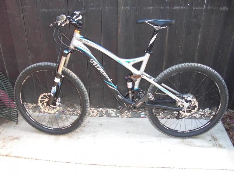 Specialized Stumpjumper FSR: A silver and black mountain bike with wide tires, positioned against a dark wooden fence. The bike features a sleek frame design and a front suspension fork, with the saddle set at a height suitable for riding. Small pebbles and a concrete surface are visible beneath the bike.