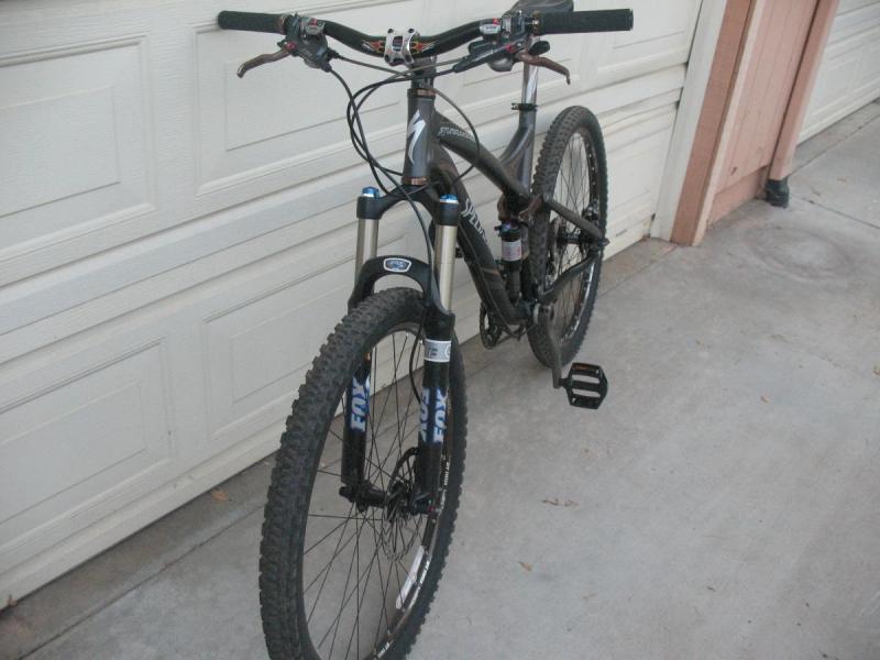 Specialized Stumpjumper FSR Elite: A mountain bike positioned at an angle against a garage door, showcasing its frame, tires, and handlebars. The bike features a black and silver color scheme, with visible suspension components and a pedal. The background includes a light-colored garage door and part of a wall.