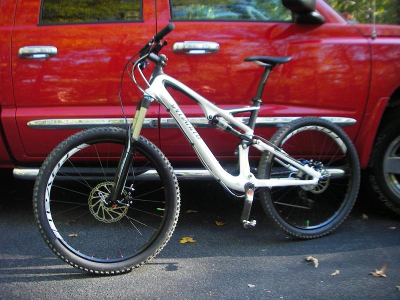 Specialized Stumpjumper: A white and black mountain bike leaning against a red pickup truck, parked on a paved surface with autumn leaves scattered around.