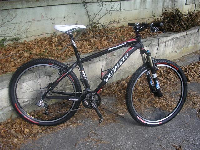 Specialized Stumpjumper: A black Specialized mountain bike resting on a path covered with dry leaves, with a concrete wall in the background. The bike features a lightweight frame, thick tires, and a white seat.