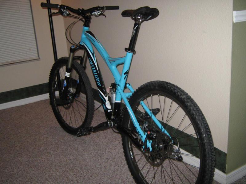 Specialized Stumpjumper: A blue and black mountain bike leaning against a wall in a room with carpeted flooring. The bike features wide tires, a sleek frame design, and a visible suspension system. There is a window in the background with blinds, and the room has a green accent wall.