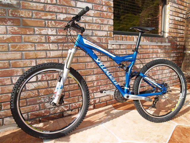 Specialized Stumpjumper: A blue mountain bike with a lightweight frame and thick, knobby tires is leaning against a brick wall. The bike features front suspension forks, a silver drivetrain, and a prominent brand logo on the frame. The background includes a window and tiles, suggesting an outdoor setting.