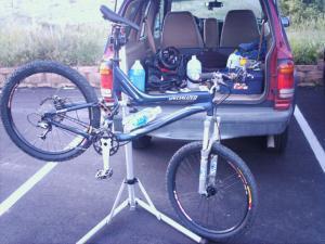 Specialized Stumpjumper: A mountain bike is being worked on while propped up on a repair stand in a parking lot. In the background, the open trunk of a vehicle reveals various items, including a water bottle and tools.