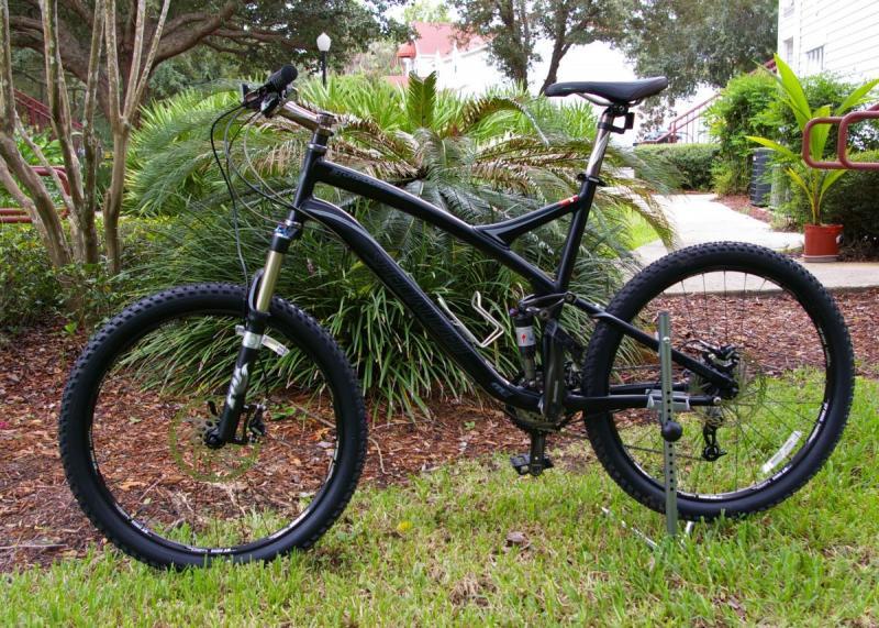Specialized Stumpjumper: A black mountain bike parked on grass, with a backdrop of lush green plants and trees. The bike features thick tires, front suspension, and a sleek frame design. The pathway in the background leads into a landscaped area, indicating a recreational setting.