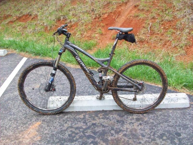 Specialized Stumpjumper: A muddy mountain bike parked on a gravel surface, with grass and a dirt hillside in the background. The bike has thick tires and a suspension system, indicating usage in rough terrain.