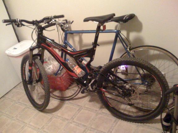 Specialized Stumpjumper: Two bicycles are parked next to each other in a room. The foreground bike is a black mountain bike with red and orange accents, while the background bike is a blue road bike. A trash bin and a red basket can be seen in the corner, and the floor features gray tiles.