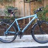 Specialized Stumpjumper: A blue bicycle with a sturdy frame and fenders, parked on a concrete surface next to a wooden fence. The bike features two seats and a compact design, suitable for commuting or casual riding.