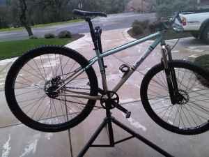 Spot Longboard: A mountain bike on a repair stand, positioned with its front wheel off the ground. The bike features a silver and teal frame, black tires with visible treads, and is set against a concrete surface with greenery in the background.