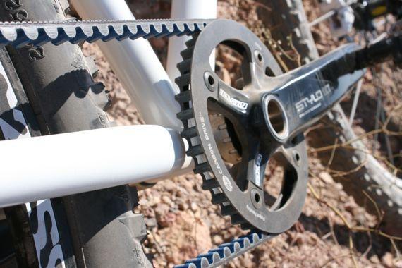 Spot Longboard: Close-up view of a bicycle's chainring and drivetrain, featuring a black crankset and blue rubber belt. The bike is positioned on rocky terrain with a white frame visible in the background.