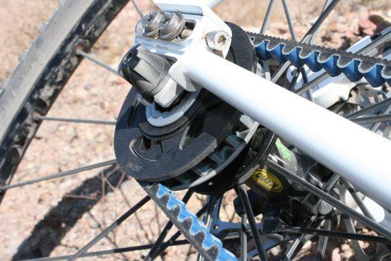 Spot Longboard: Detailed close-up of a bicycle's rear hub, showcasing the drive system with a black cog, blue belt, and a white frame. The background features a rocky outdoor terrain.
