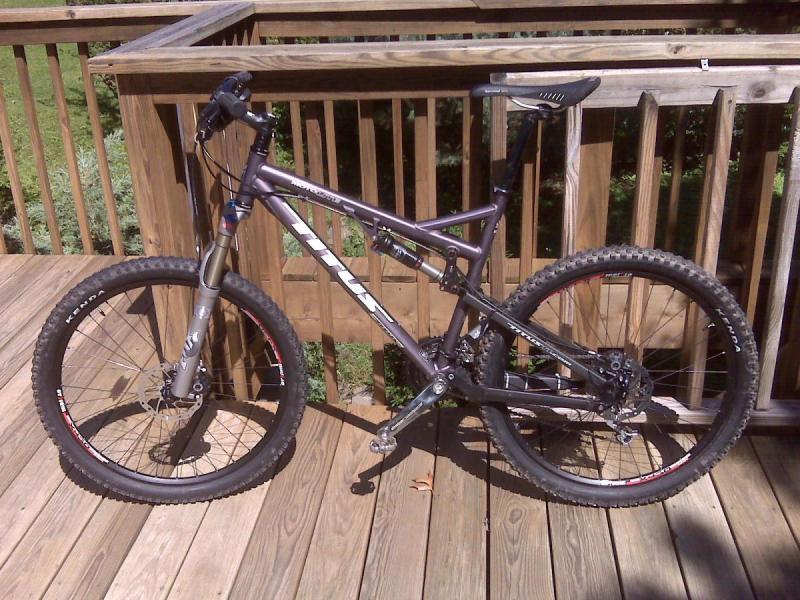 Titus Moto Lite: A purple mountain bike with thick tires and a dual-suspension frame is positioned on a wooden deck. The bike features a sleek design, front suspension forks, and visible gear mechanisms, set against a background of green foliage.