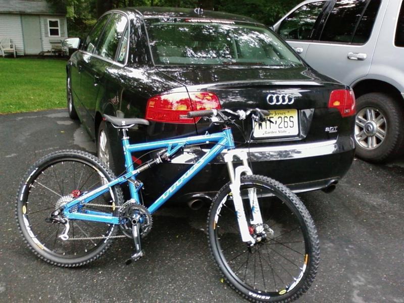 Titus Racer X: A black Audi sedan parked in a driveway next to a mountain bike with blue and white colors. The bike is leaning against the rear of the car. The background features greenery and a small house.