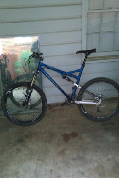 Titus Racer X: A blue mountain bike with a silver frame is leaning against a wall on a porch, with a mirror reflecting the outdoors visible in the background. The ground appears to be a concrete surface.