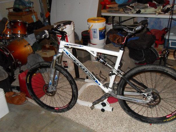 Titus Racer X: A mountain bike with a white frame and blue accents is parked on a carpeted floor in a cluttered garage. Various items, including a drum set and paint buckets, are visible in the background.