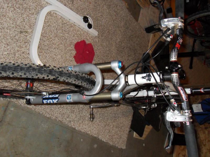 Titus Racer X: A top-down view of a mountain bike resting on a carpeted surface. The bike features thick, knobby tires, a lightweight aluminum frame, and front suspension components. Several colorful components like the handlebars and disc brakes are visible, along with a red cloth in the background and tools or bike accessories scattered around.