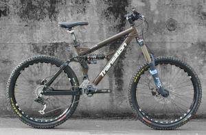 Transition Preston FR: A brown mountain bike with sturdy tires and a sleek design, positioned against a textured gray wall. The bike features a front suspension fork and a prominent brand logo on the frame.