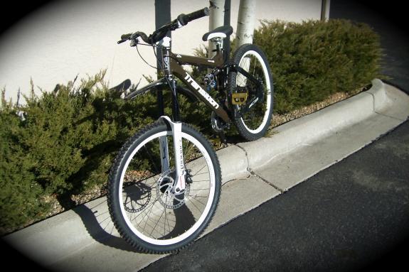 Transition Preston FR: A mountain bike with a brown frame and white tires is leaned against a curb next to some shrubs. The bike features disc brakes and prominent handlebars, positioned on a paved surface with a light-colored wall in the background.