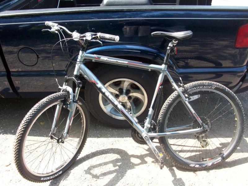 Trek 3500: A silver mountain bike leaned against a dark blue pickup truck, showing the bike's frame, handlebars, and front wheel with knobby tires. The pickup truck's rear tire is visible in the background, and the scene is set outdoors on a gravel surface.