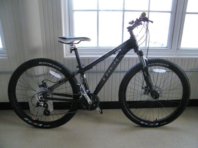 Trek 3700: A black mountain bike with wide tires, positioned indoors, next to a window. The bike features a sleek frame with visible branding, a comfortable saddle, and a front fork designed for stability. The background includes a neutral-colored floor and white window frames.