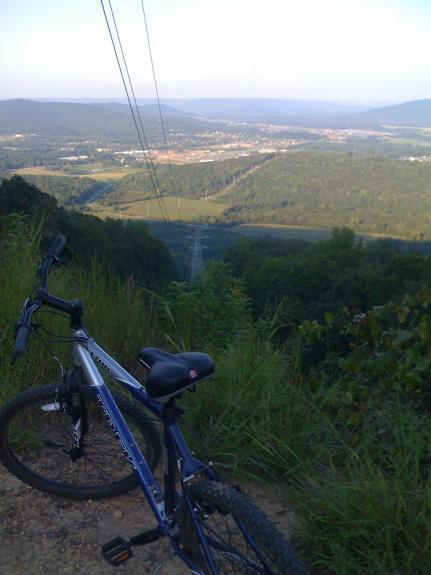 Trek 3700: A mountain bike rests on a dirt path overlooking a scenic valley filled with greenery and rolling hills, with power lines running across the sky and a distant town visible in the valley below.