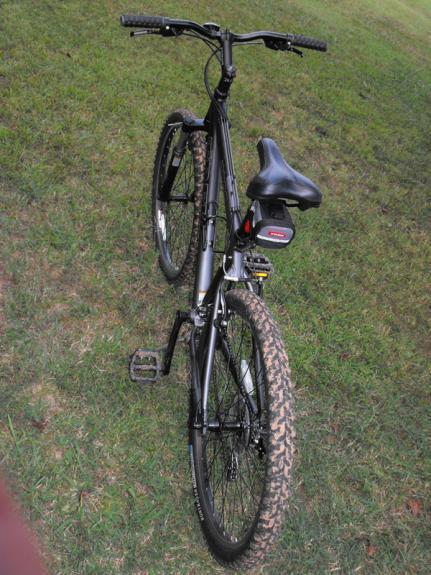 Trek 3700: A black mountain bike positioned on grassy terrain, showing the seat, handlebars, and rear wheel. The bike features a rugged design with knobby tires suitable for off-road rides.