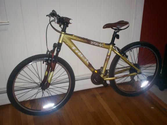 Trek 3700: A mountain bike with a gold and black frame, featuring a brown saddle and reflective tires, parked indoors on a hardwood floor. The bike has front suspension and visible gears, with a plain white wall and a red accent wall in the background.