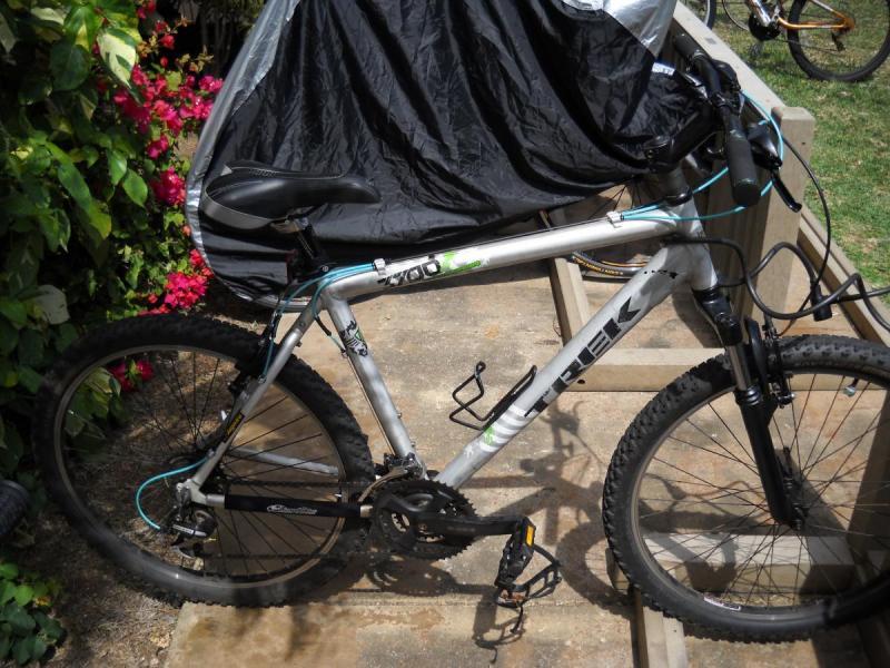 Trek 3700: A silver Trek mountain bike parked on a concrete surface, partially covered with a black protective cover. Colorful flowers and green foliage are visible in the background, creating a vibrant outdoor scene.