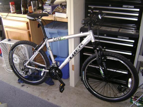 Trek 3700: A silver Trek mountain bike standing on a carpeted floor in a garage. The bike features black tires, a sturdy frame, and various gear components visible. In the background, there are wooden cabinets and a tool storage unit. A fan is positioned nearby, and various items are scattered on the work surface.
