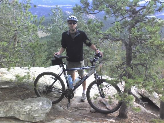Trek 3700: A person wearing a helmet and athletic clothing stands next to a mountain bike on a rocky outcrop surrounded by trees. The background features rolling hills and distant mountains under a partly cloudy sky.