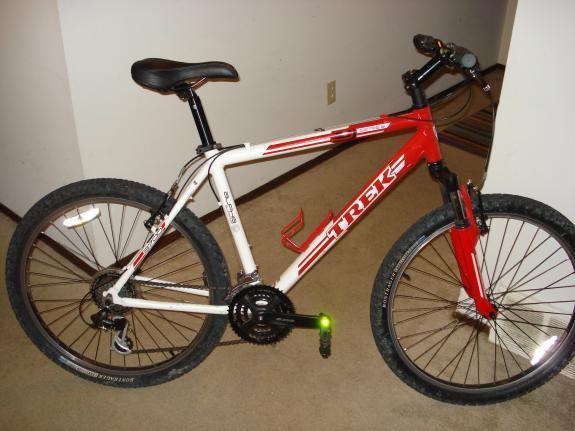 Trek 3700: Alt tag: A red and white Trek mountain bike leaning against a wall, with knobby tires and a basic design, sitting on a carpeted floor.